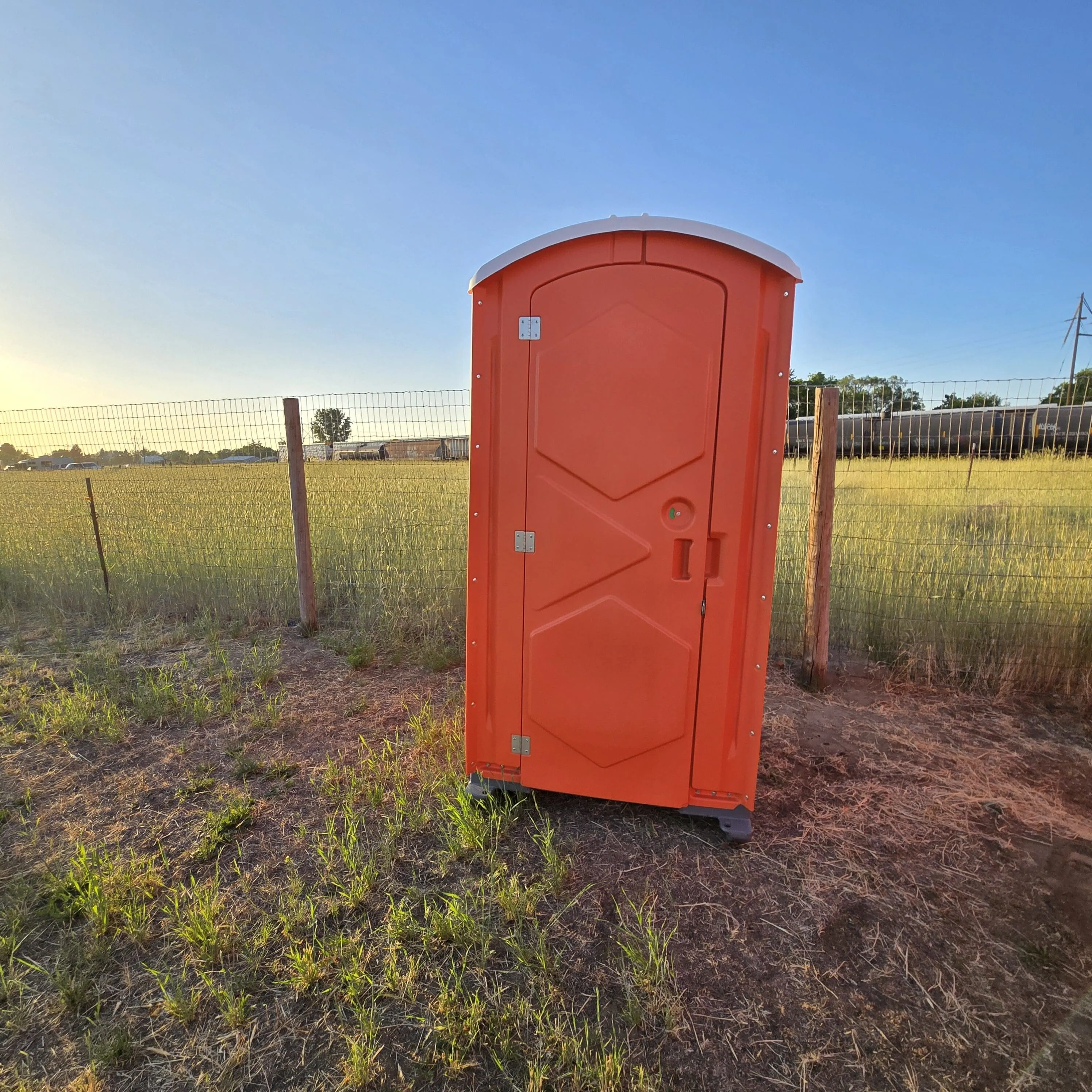 A single orange Garcia Brothers portable toilet standing in an open field at golden hour